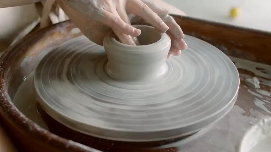 Two hands pulling the wall of a small pot up on a turning wheel.